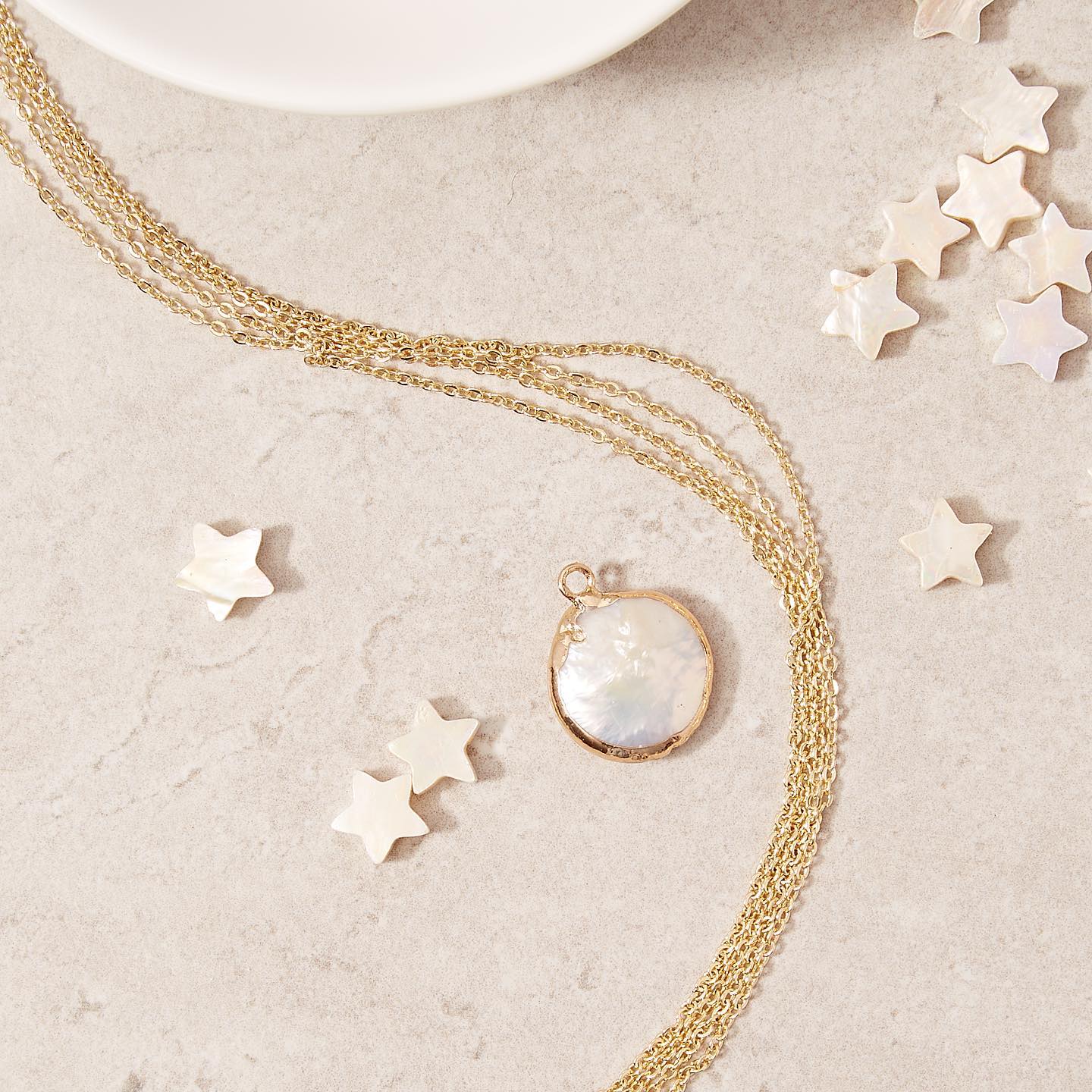 A picture of a pearl pendant on sand colored ground with star beads surrounding it and a gold chain