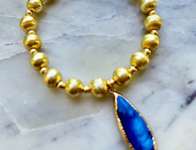 A picture of a gold beaded bracelet with a gold lined royal blue teardrop pendant