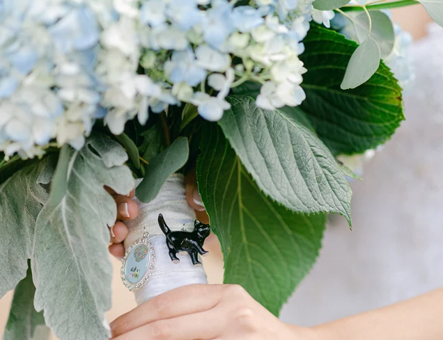 Picture of a woman at a wedding holding a bouquet of flowers; zoomed into a picture of a small black cat charm and a regency charm on the bouquet stem.